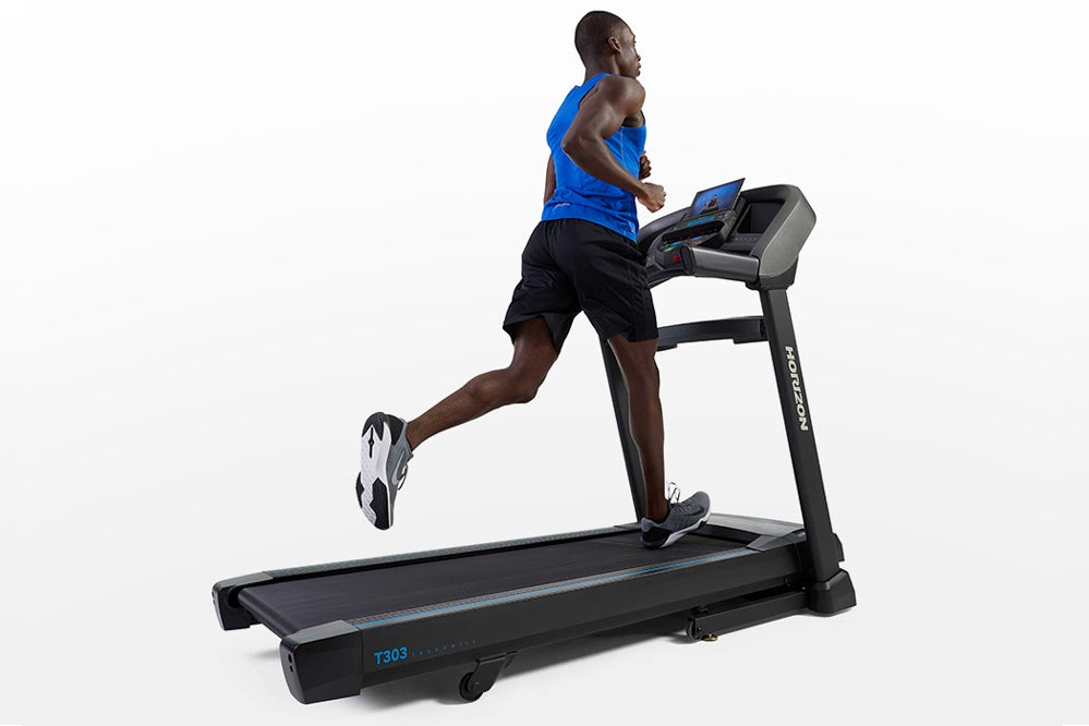Man running on black Horizon T303 folding treadmill with blue digital display, wearing blue tank top and black shorts on whit
