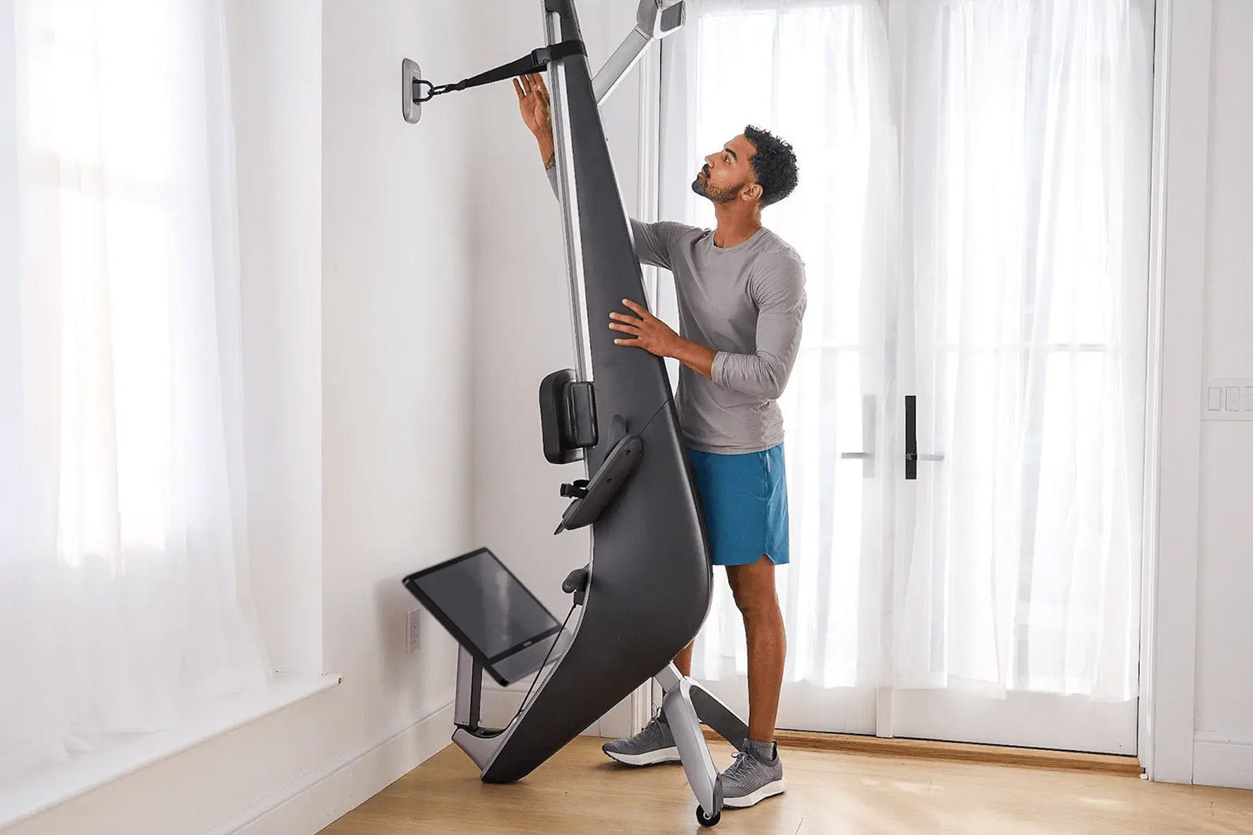Man using black and silver Hydrow Arc Rower in home gym with large windows, wearing gray shirt and blue shorts