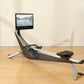 Hydrow Arc Rower with black seat and silver frame, mounted touchscreen displaying water rowing video, on wooden floor in home