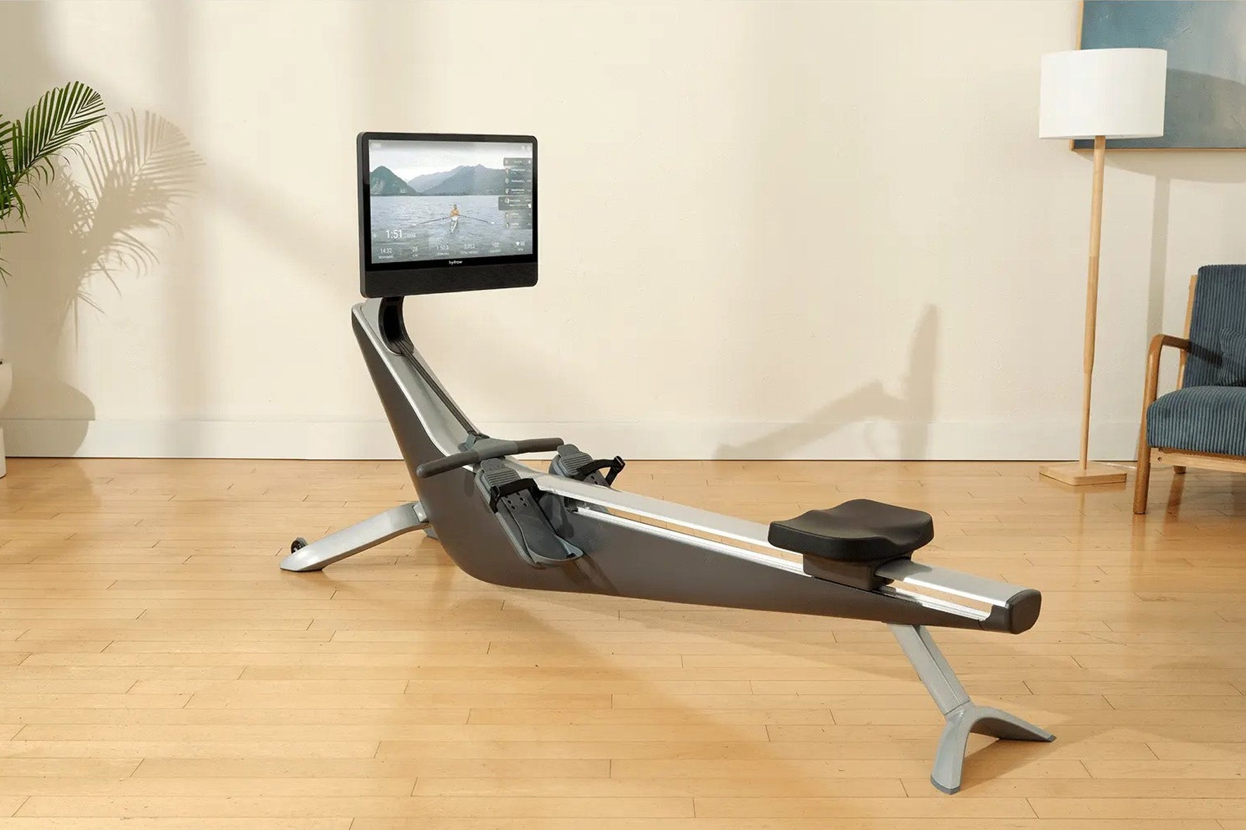 Hydrow Arc Rower with black seat and silver frame, mounted touchscreen displaying water rowing video, on wooden floor in home