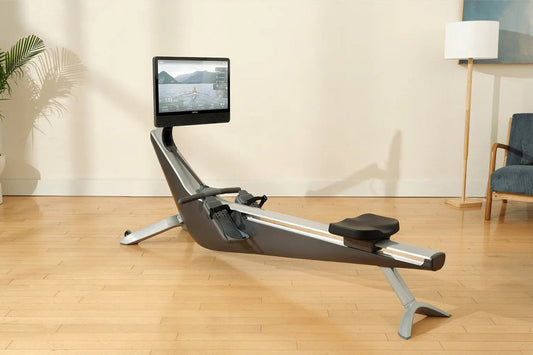 Hydrow Arc Rower with black seat and silver frame, mounted touchscreen displaying water rowing video, on wooden floor in home