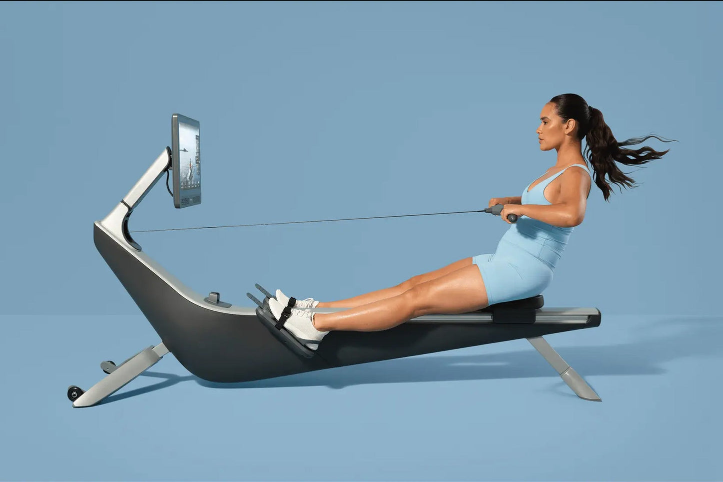 Woman in light blue athletic wear using black and silver rowing machine with attached digital screen on blue background