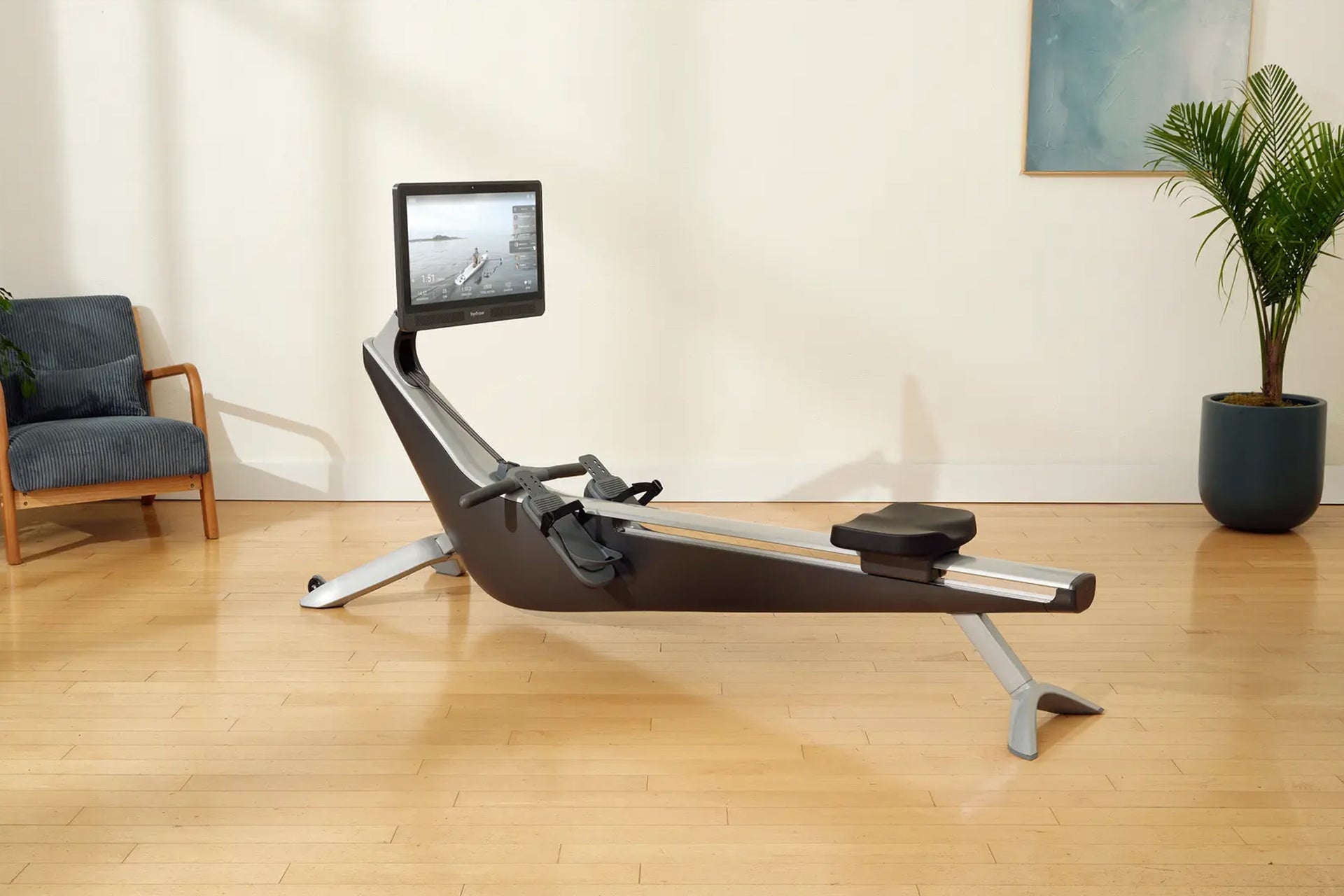 Black and silver rowing machine with attached digital screen displaying water scene, positioned on wooden floor in home inter