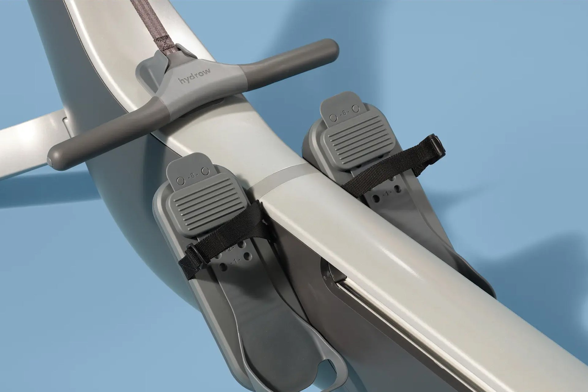 White Hydrow Origin rowing machine with gray handles and two gray footrest pedals with black straps on blue background