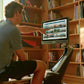 Man using Hydrow Pro rowing machine with touchscreen display showing scenic water images, positioned in front of wooden books