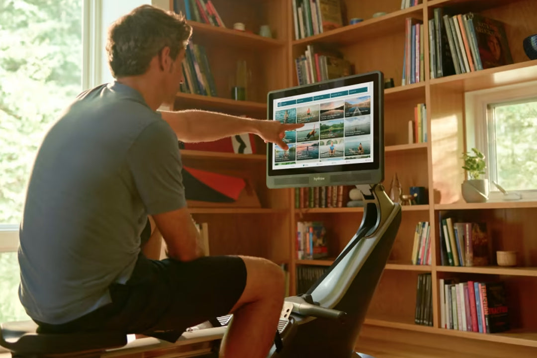 Man using Hydrow Pro rowing machine with touchscreen display showing scenic water images, positioned in front of wooden books