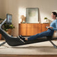 Woman in blue long-sleeve shirt and navy leggings using black Hydrow Pro rowing machine in home gym setting with wood dresser