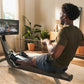 Man using black rowing machine with digital monitor displaying workout content in home gym setting