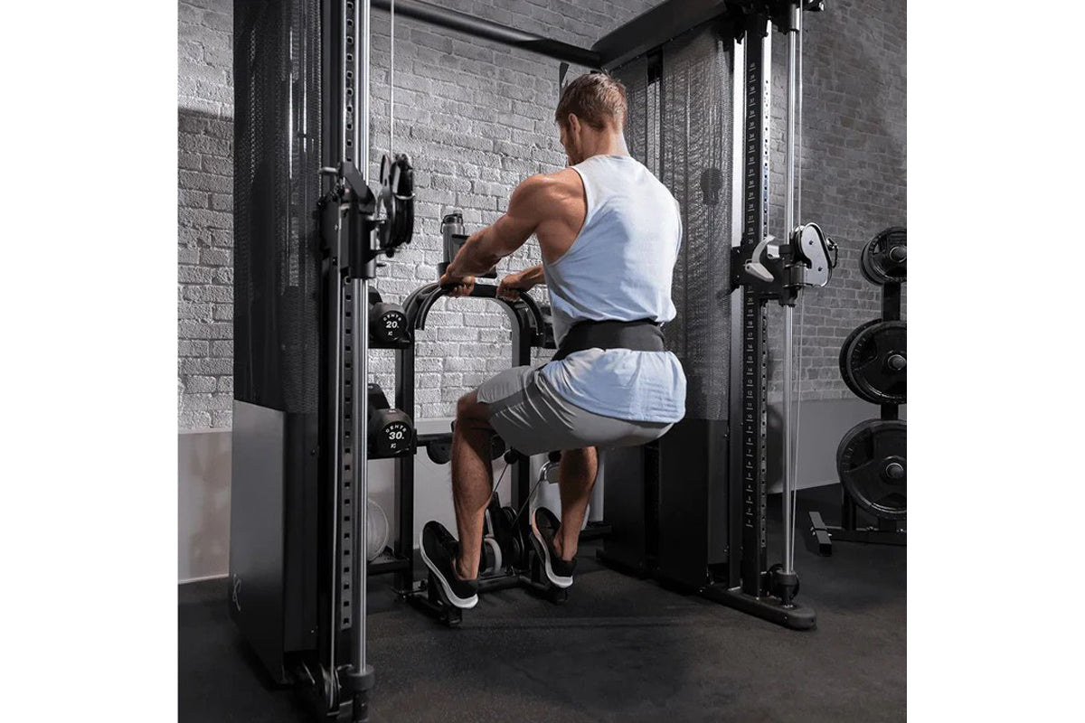 Man using black Smith functional trainer machine with cable attachments in gym setting, wearing white tank top and gray short