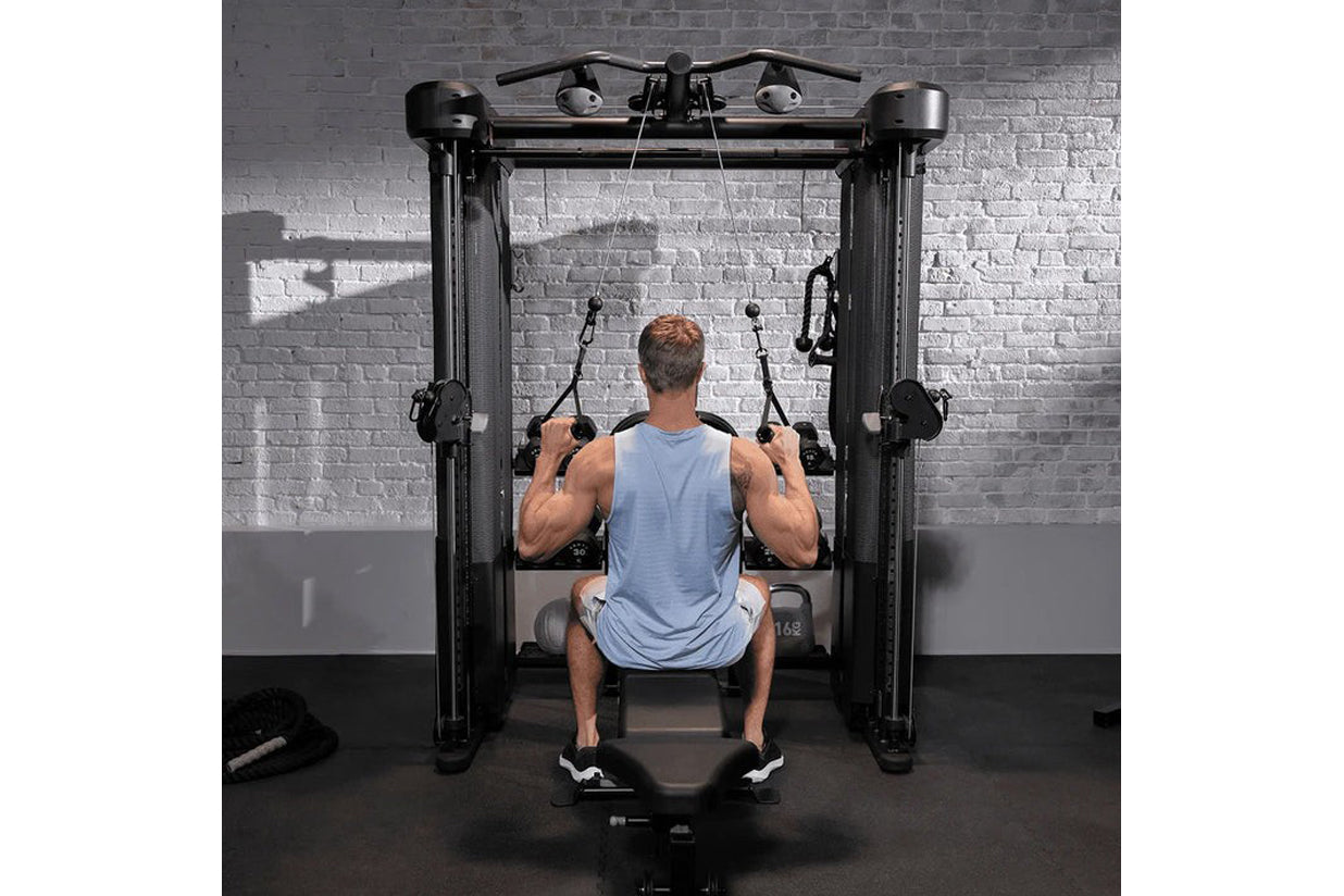 Man using black dual-column functional trainer machine with cable handles in gym setting against brick wall