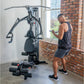 Black multi-gym system with cable pulley, seat, and foot rest, in use by man in gym setting with brick wall