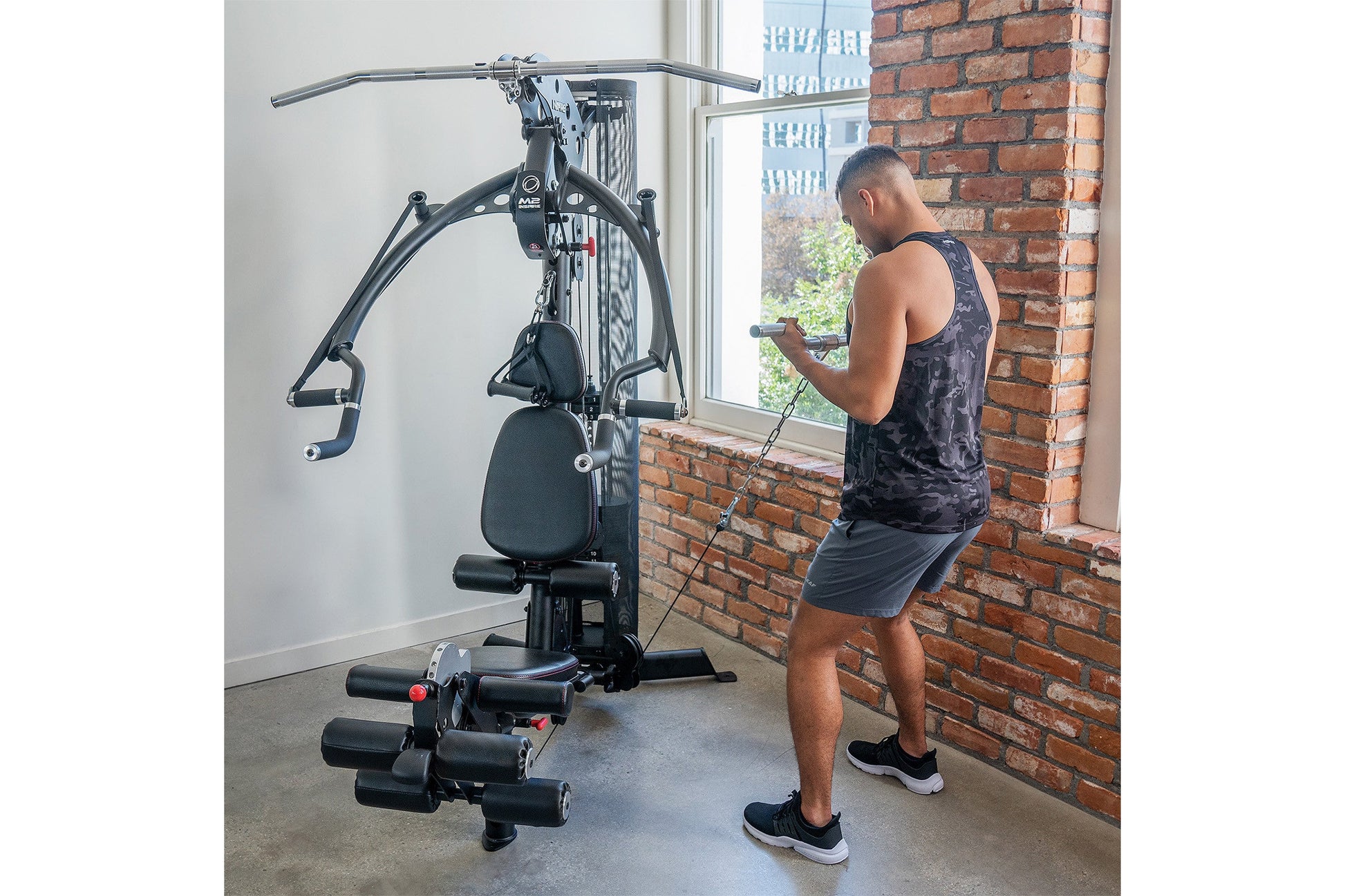 Black multi-gym system with cable pulley, seat, and foot rest, in use by man in gym setting with brick wall