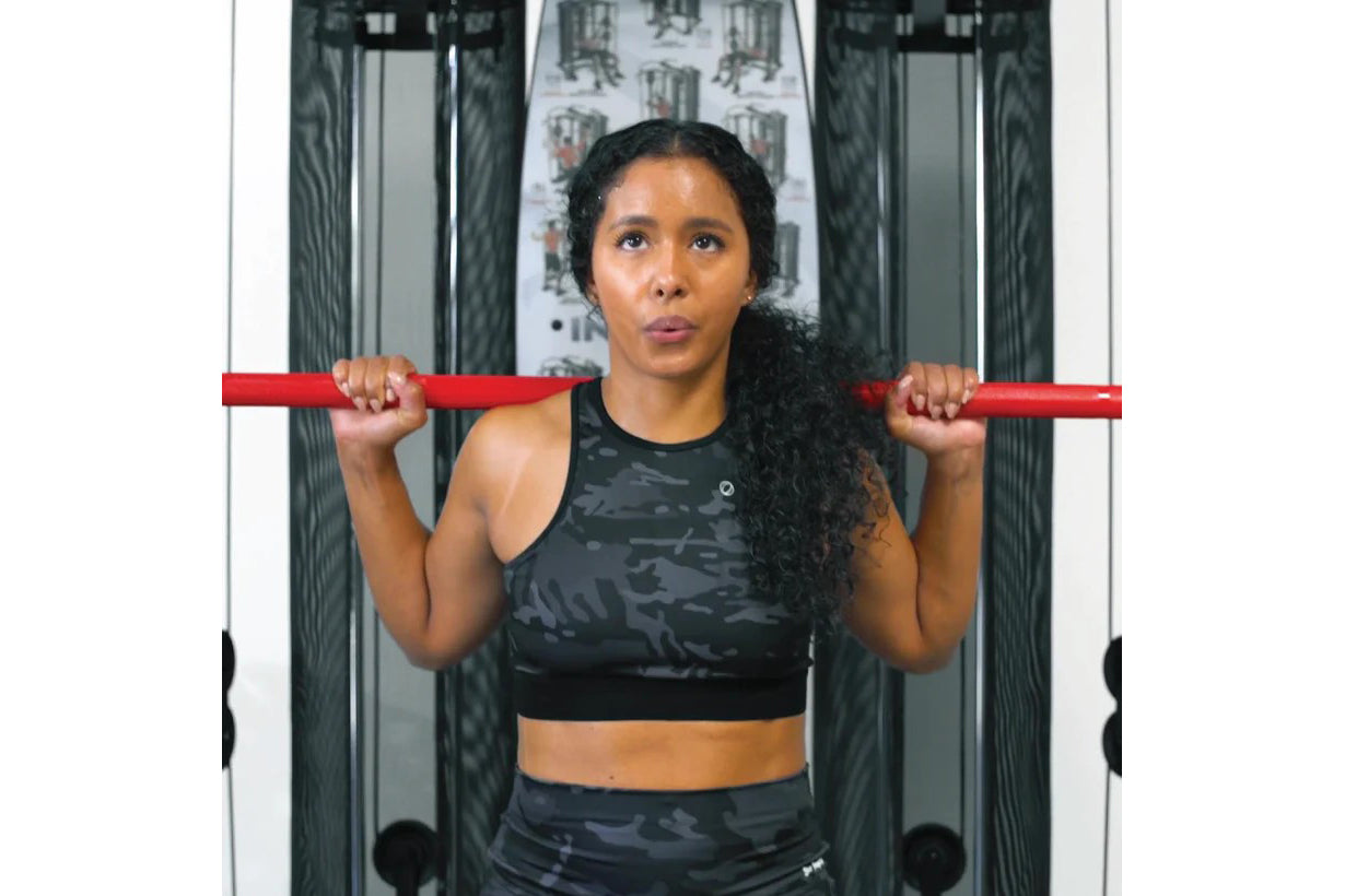 Woman wearing black camo sports bra and leggings holding red barbell across shoulders in gym setting