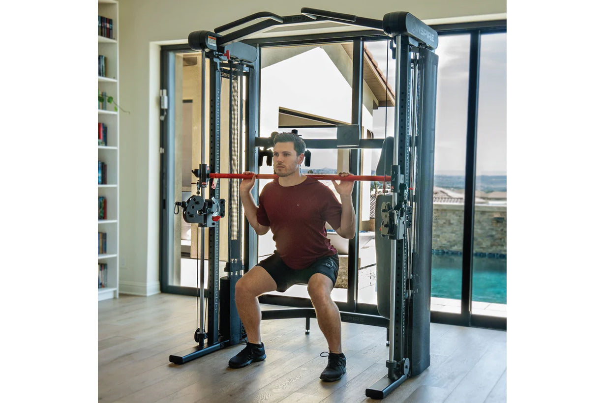 Man in burgundy t-shirt and black shorts using black Smith functional trainer with red bar in modern home gym with ocean view