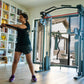 Woman using blue and red Smith functional trainer machine with resistance cables in home gym setting with bookshelves