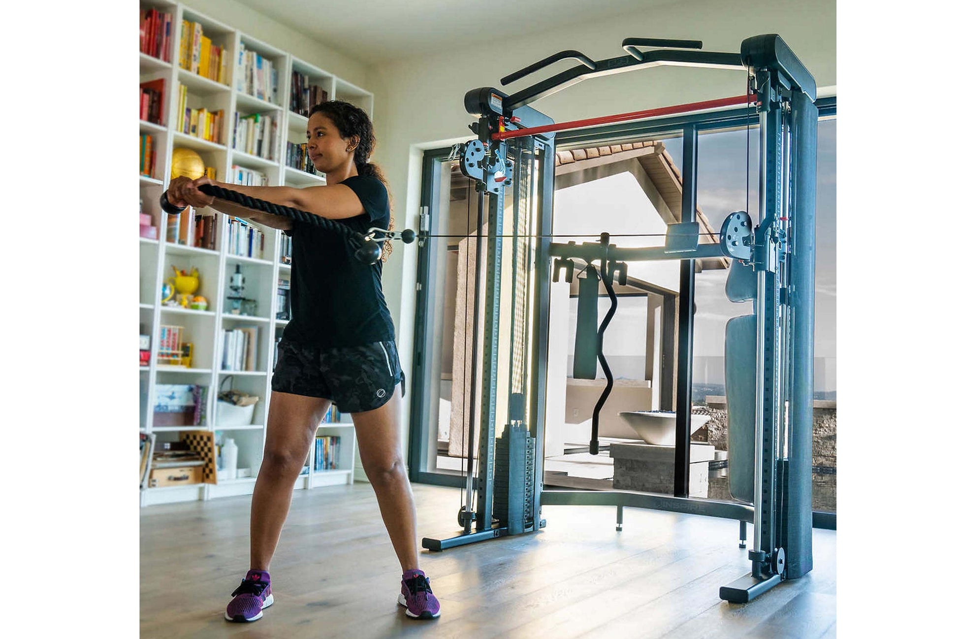 Woman using blue and red Smith functional trainer machine with resistance cables in home gym setting with bookshelves