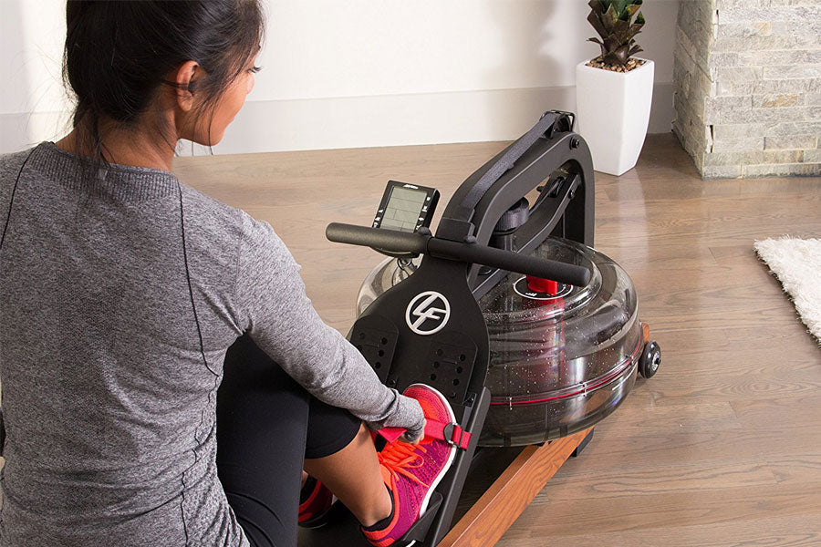 Woman using black water rower with digital display on wooden floor, wearing gray top and pink sneakers