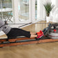 Woman using wooden water rower with black frame and clear water tank on hardwood floor, indoors