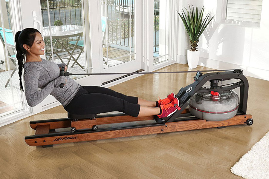 Woman using wooden water rower with black frame and clear water tank on hardwood floor, indoors