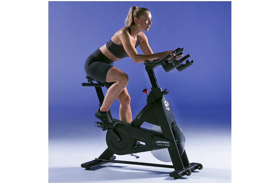 Woman in black sports bra and shorts riding Life Fitness IC3 indoor stationary bike against blue background