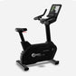 Black upright exercise bike with digital touchscreen display, padded seat, handlebars, and foot pedals on gray background