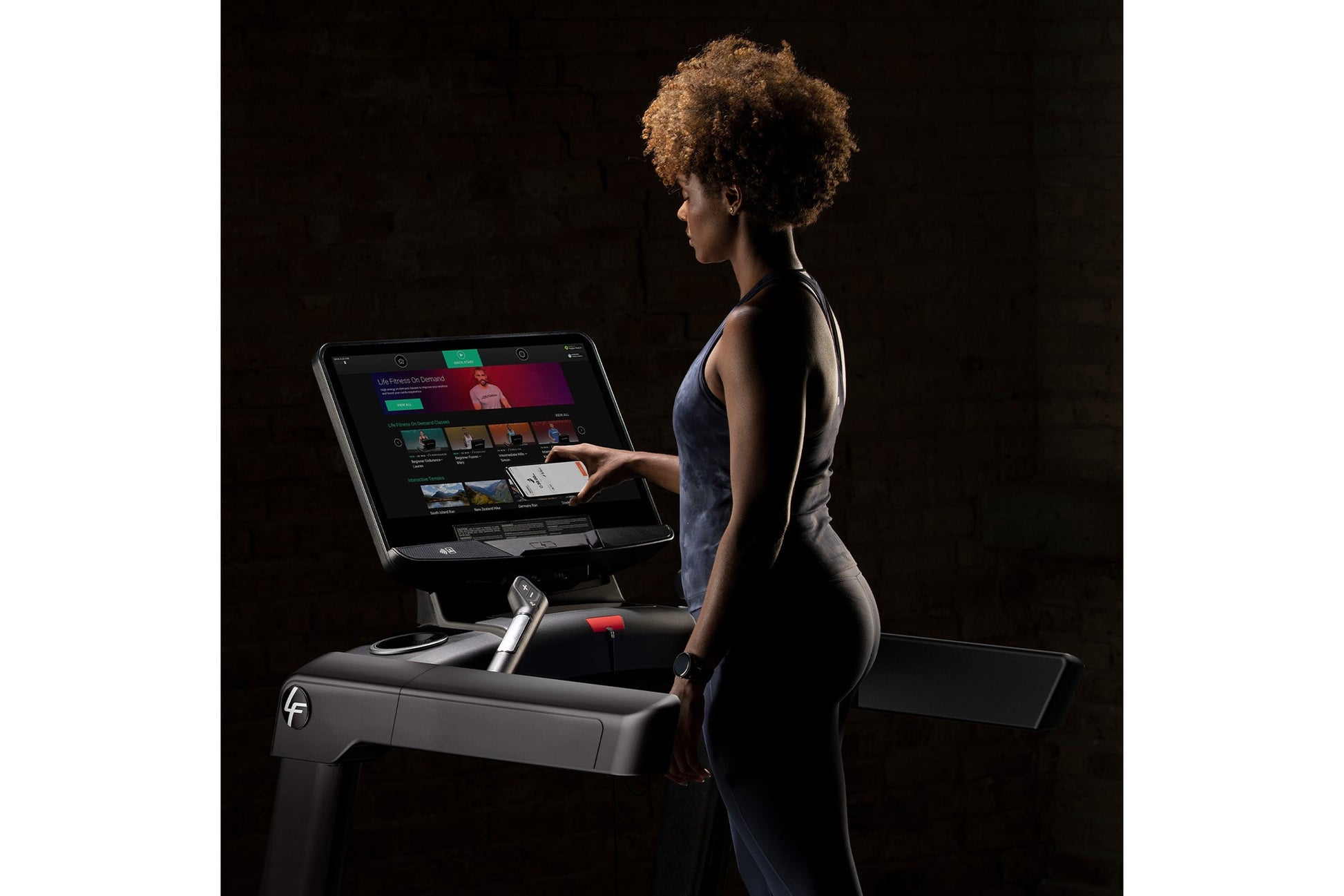 Woman wearing dark blue athletic tank top using Life Fitness Integrity+ treadmill with digital touchscreen display against bl