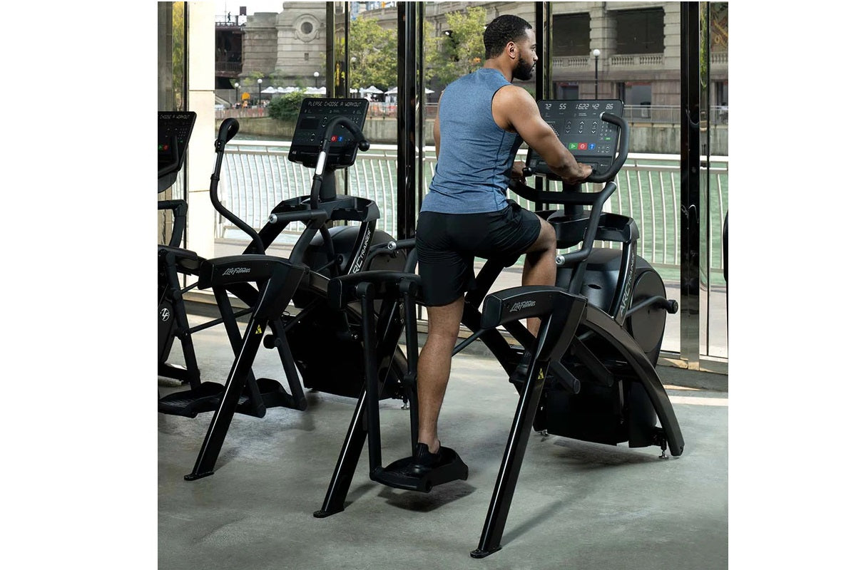 Man wearing blue sleeveless tank top using black Life Fitness arc trainer elliptical machine in gym with city view windows