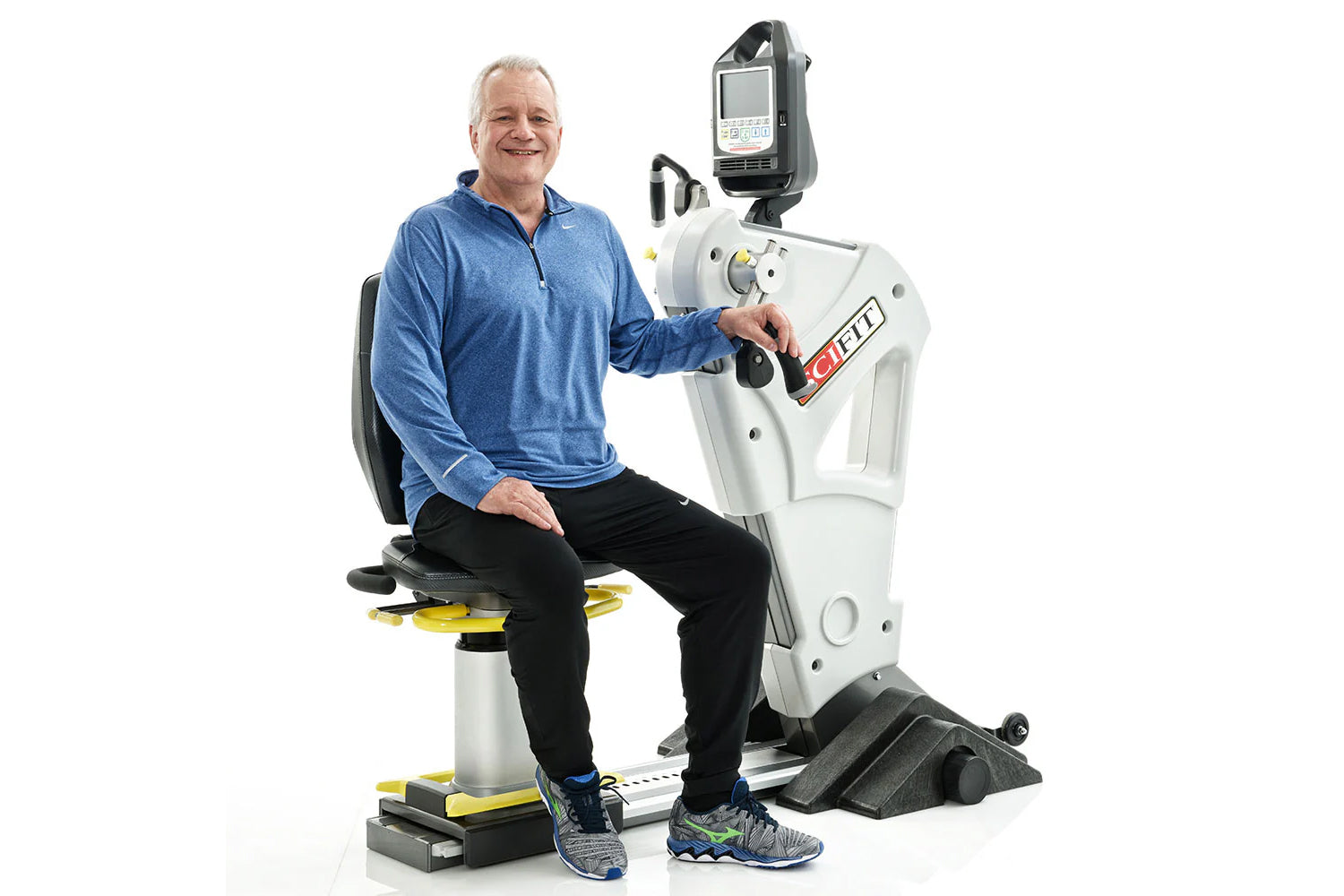 Man in blue jacket seated on white and yellow exercise machine with digital display monitor, white background