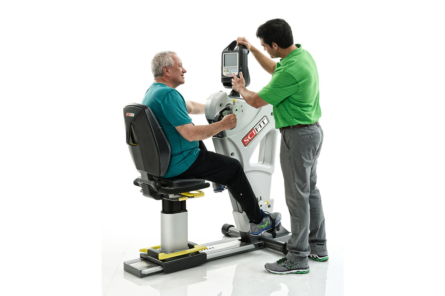 Older man in teal shirt seated on white and black exercise machine with trainer in green shirt adjusting digital monitor abov