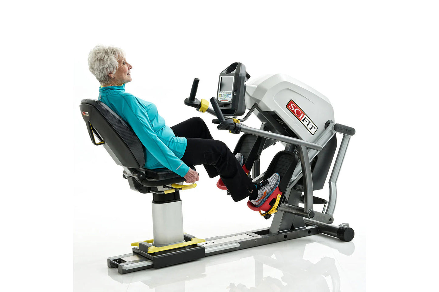 Older woman in turquoise jacket using white and black SCIFIT recumbent stepper with digital display, handlebars and foot peda