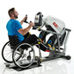 Man in blue shirt using white and black recumbent stepper exercise machine with digital display, seated in wheelchair on whit