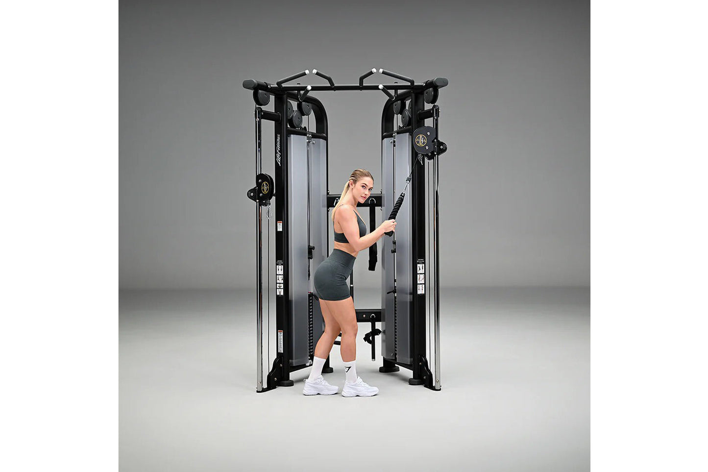 Black and silver dual adjustable pulley machine with woman in black sports bra and shorts using cable attachment, on gray bac