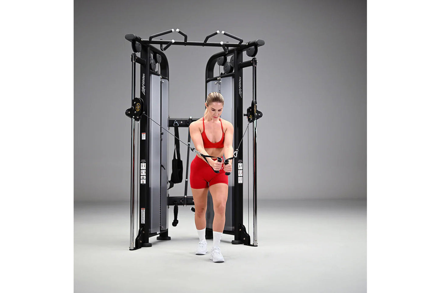 Woman in red sports bra and shorts using black and silver dual adjustable pulley machine on gray background