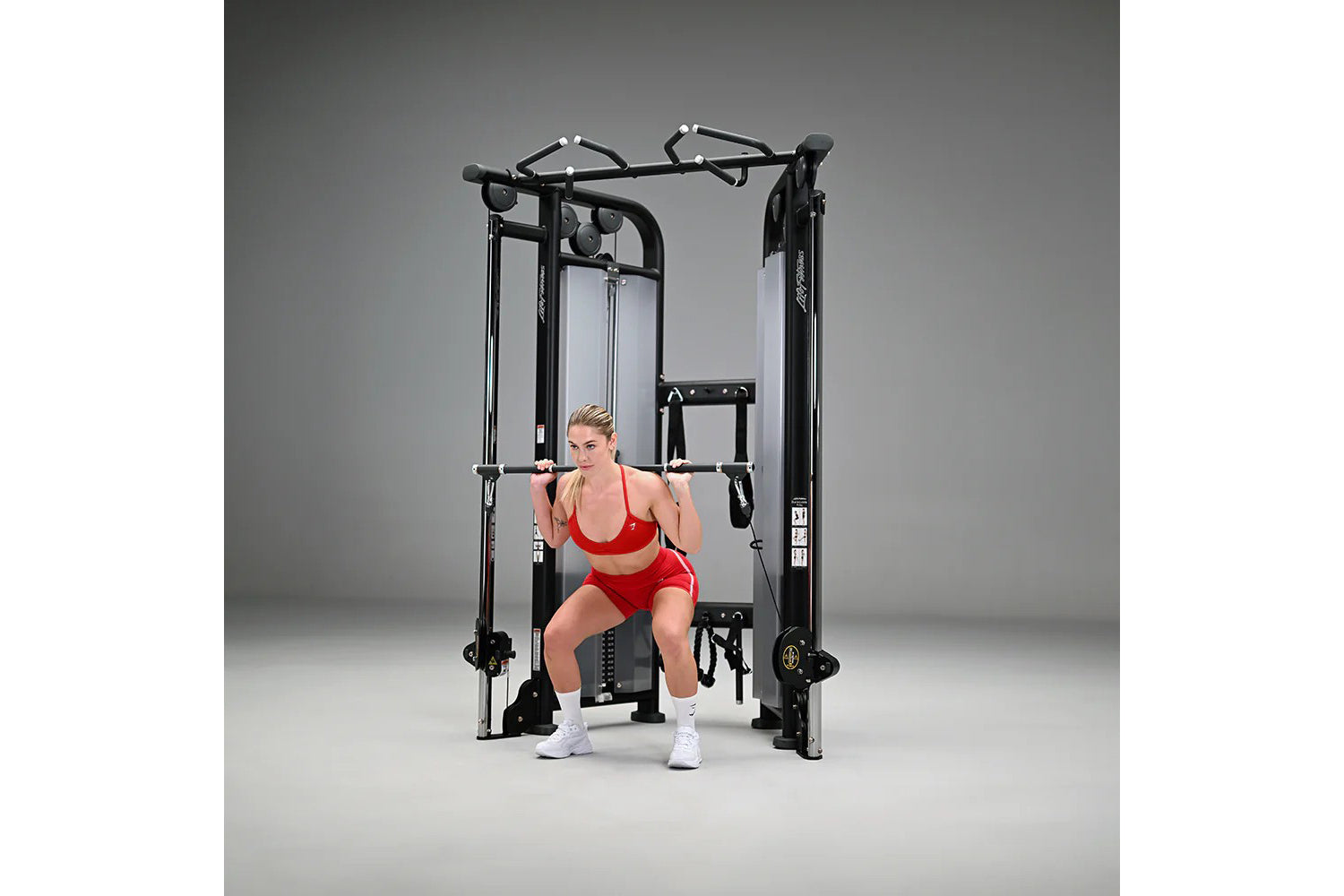 Woman in red sports bra and shorts using black dual adjustable pulley cable machine on gray background