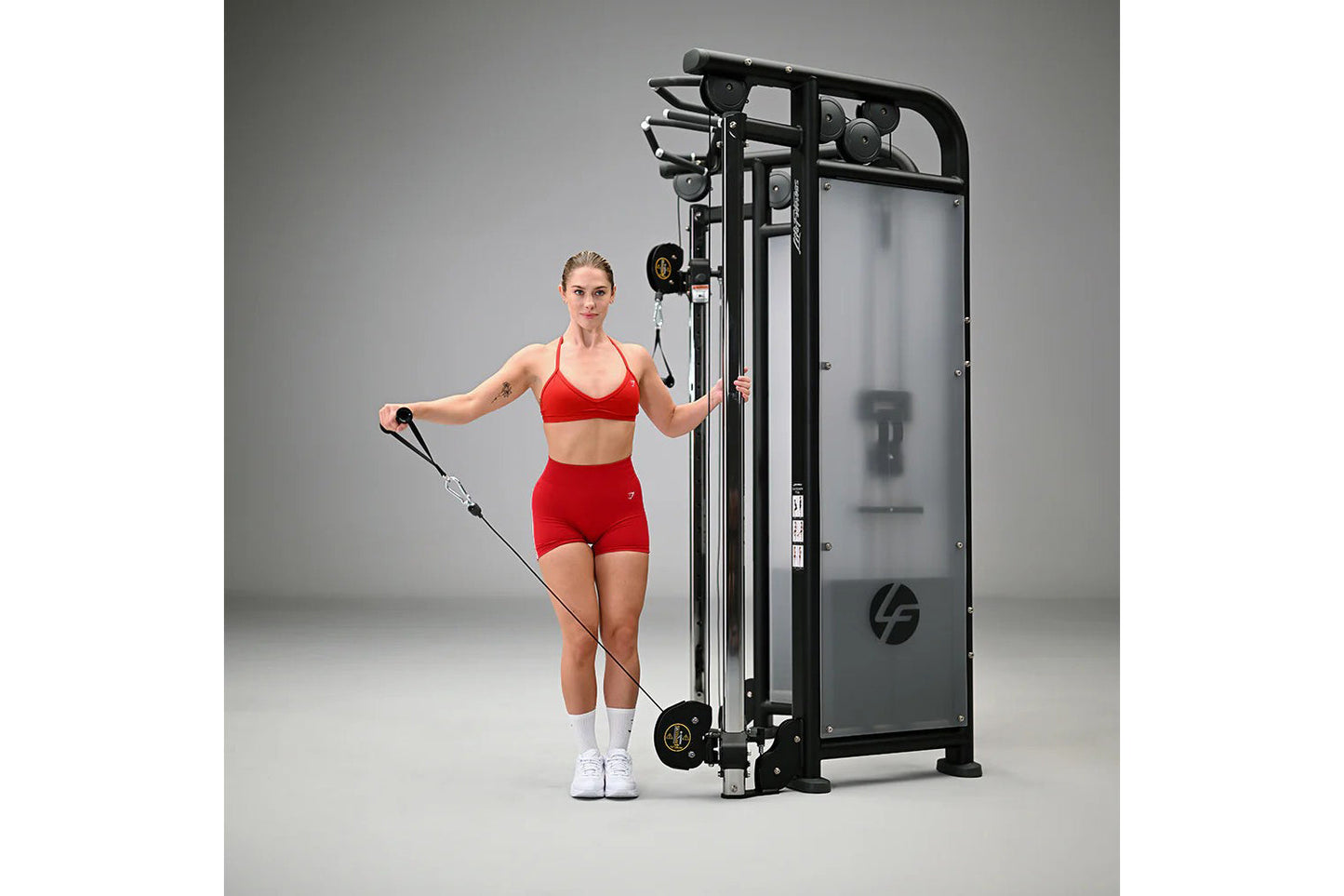 Woman in red sports bra and shorts using black dual adjustable pulley machine with cable handles on gray background
