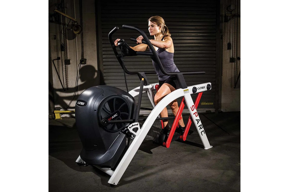 Woman using red and white arc trainer elliptical with black fan wheel in gym setting, wearing purple tank top and black short
