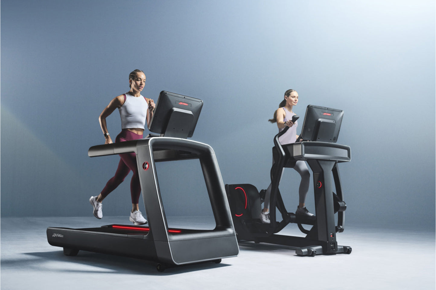 Two models demonstrating Life Fitness Symbio incline elliptical machines with black frames, digital screens, and red accent d