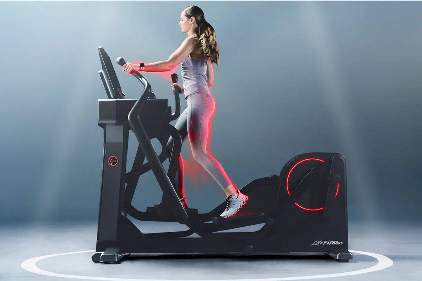 Woman using black Life Fitness elliptical with red accent lighting and digital display, worn by model on blue background
