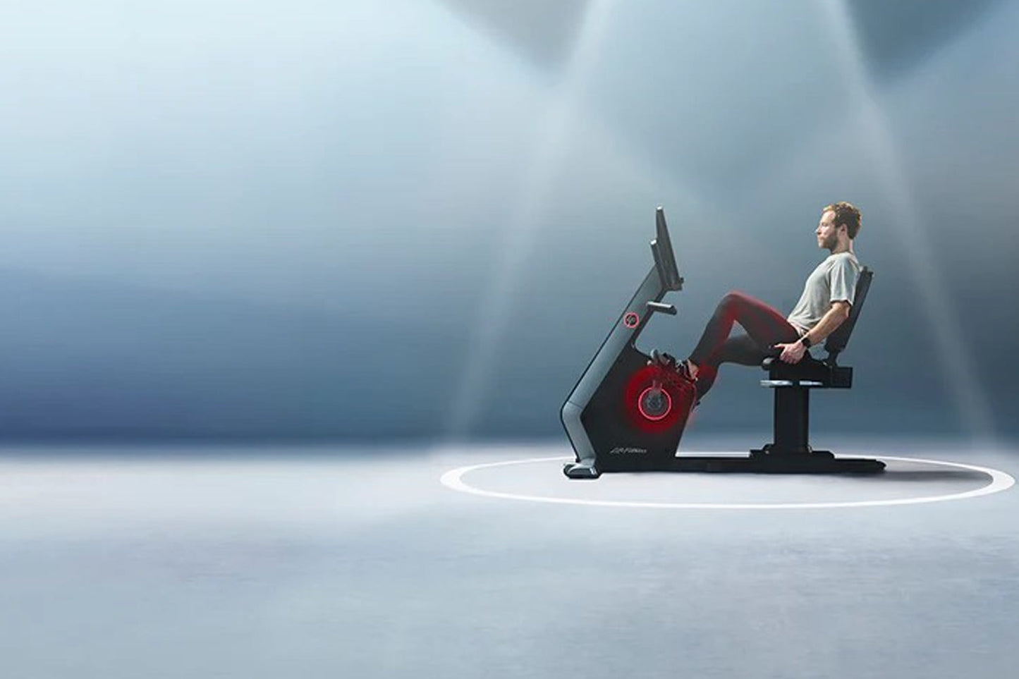 Man wearing beige t-shirt and burgundy pants seated on Life Fitness Symbio red and black recumbent exercise bike against blue