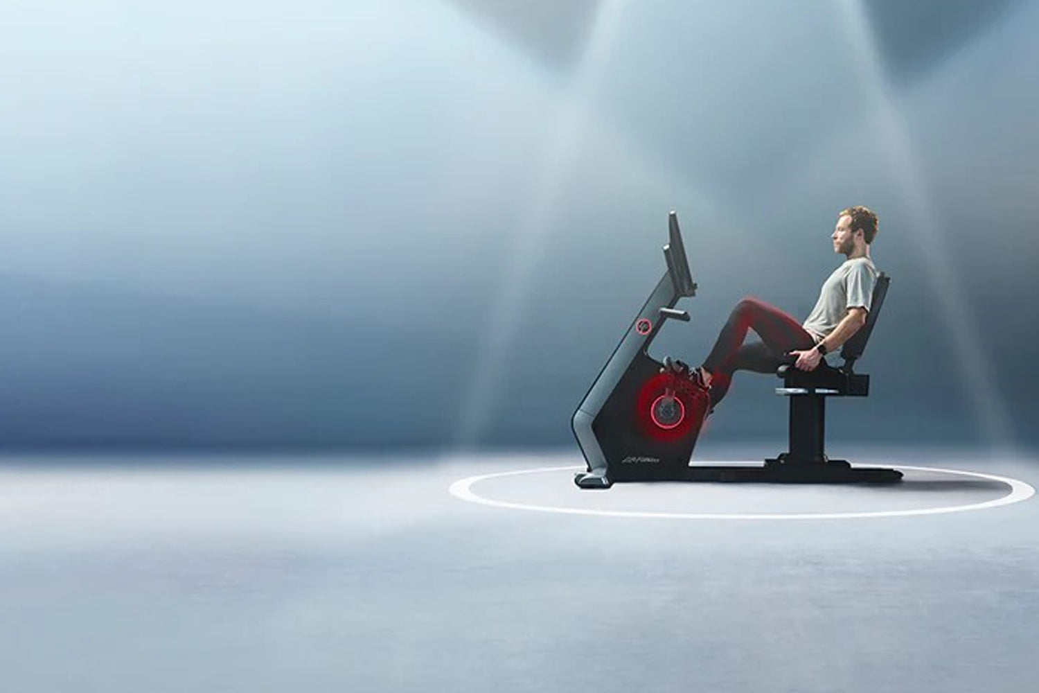 Man wearing beige t-shirt and burgundy pants seated on Life Fitness Symbio red and black recumbent exercise bike against blue