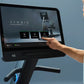 Black recumbent exercise bike with touchscreen display showing Symbio interface, user's hand touching screen, blue background