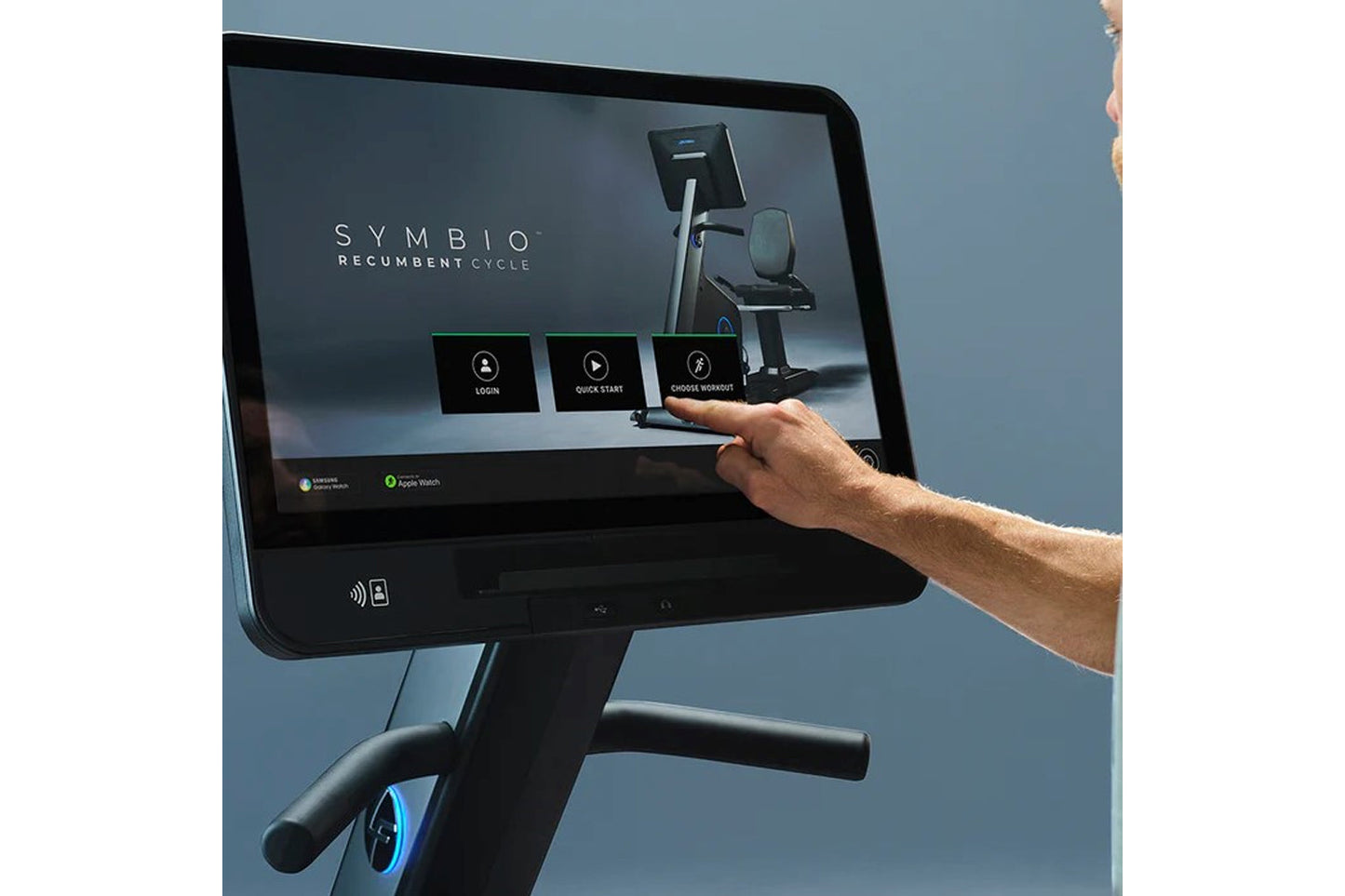 Black recumbent exercise bike with touchscreen display showing Symbio interface, user's hand touching screen, blue background