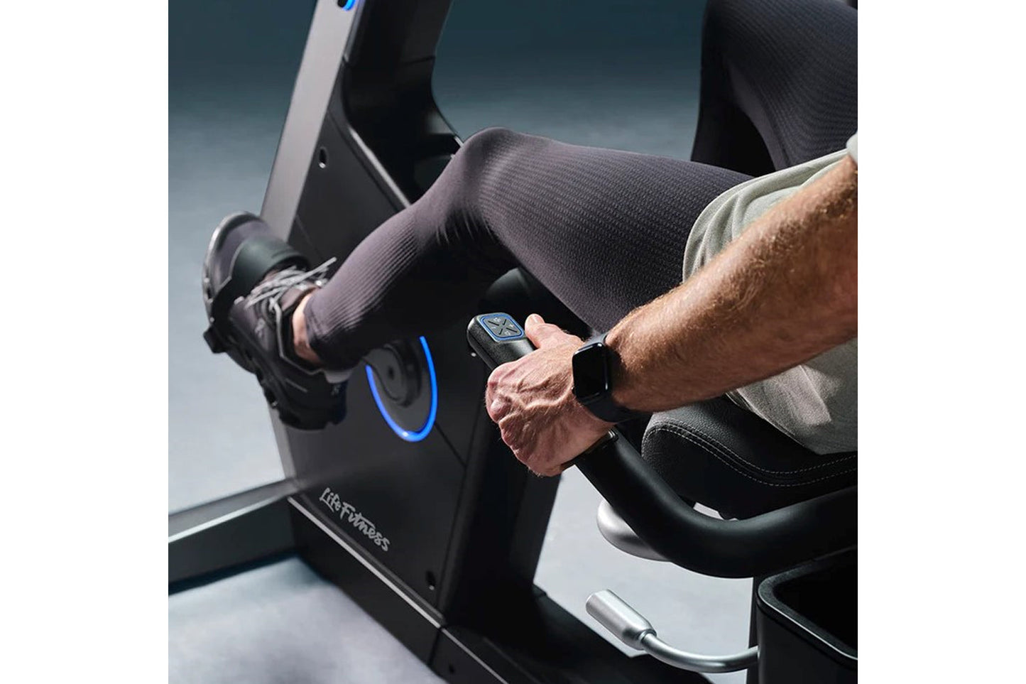 Man wearing smartwatch and dark compression tights on Life Fitness Symbio recumbent exercise bike with blue accent lighting