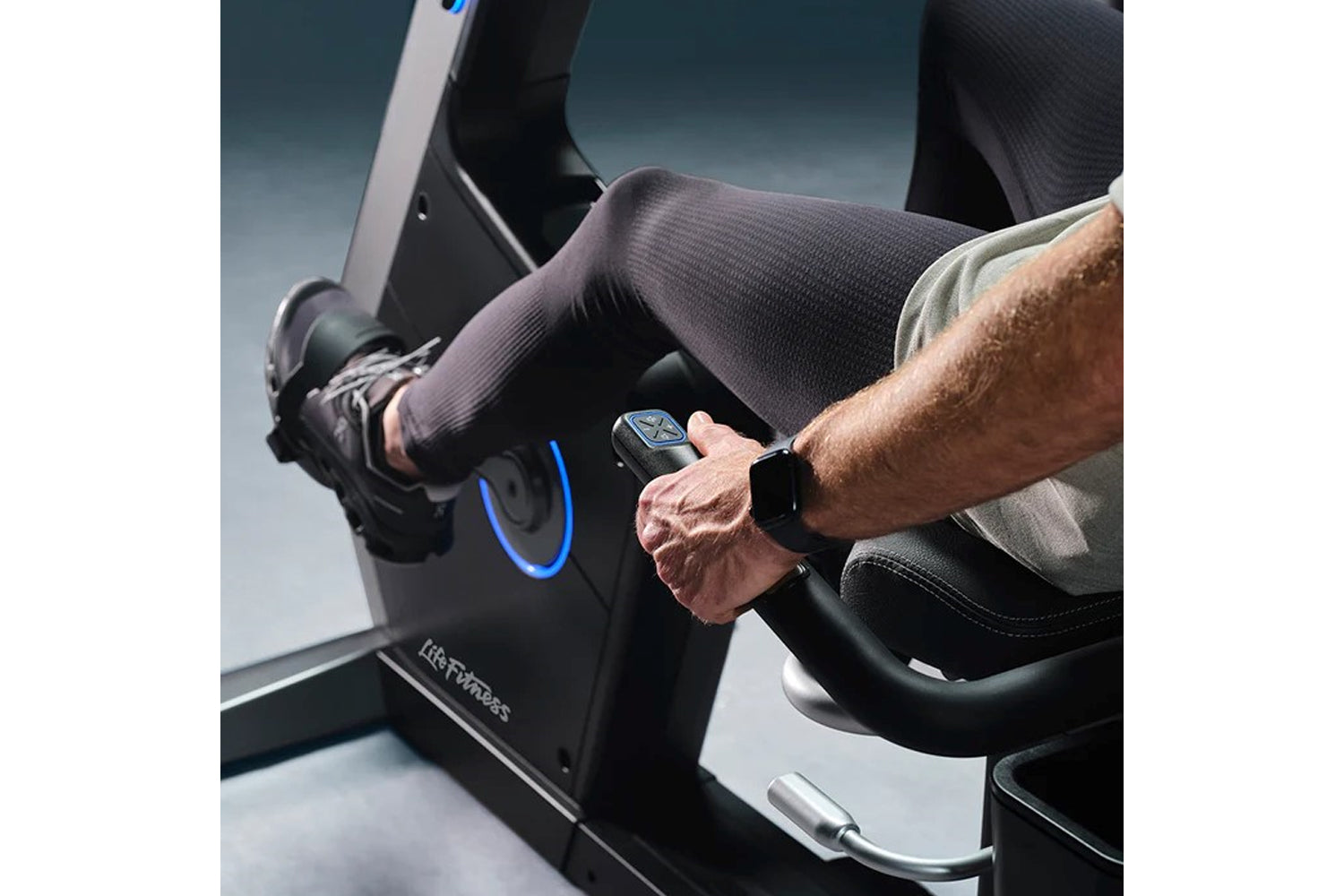 Man wearing smartwatch and dark compression tights on Life Fitness Symbio recumbent exercise bike with blue accent lighting