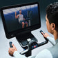 Person using Life Fitness Symbio Switch stationary exercise bike with touchscreen display showing workout video and handheld 