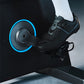 Black and gray indoor cycling shoe with blue accent straps on stationary bike pedal with blue-rimmed flywheel, worn by model 