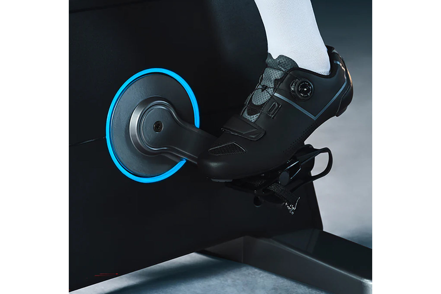 Black and gray indoor cycling shoe with blue accent straps on stationary bike pedal with blue-rimmed flywheel, worn by model 