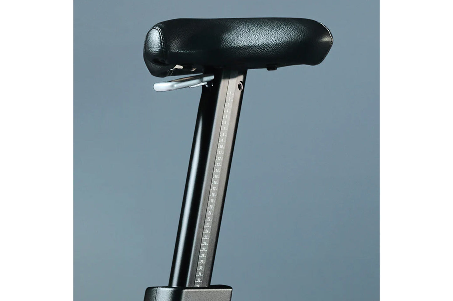 Stationary bike seat with black padded top, metal post with height adjustment markings, and gray background