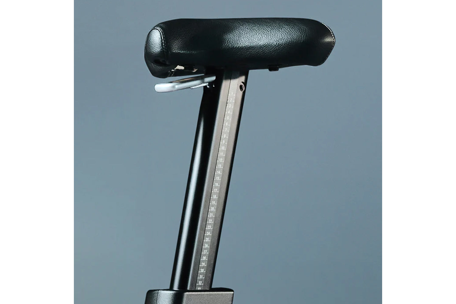 Stationary bike seat with black padded top, metal post with height adjustment markings, and gray background