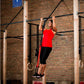 Woman wearing red tank top and black capris using red pull-up assist band on black pull-up bar in gym setting